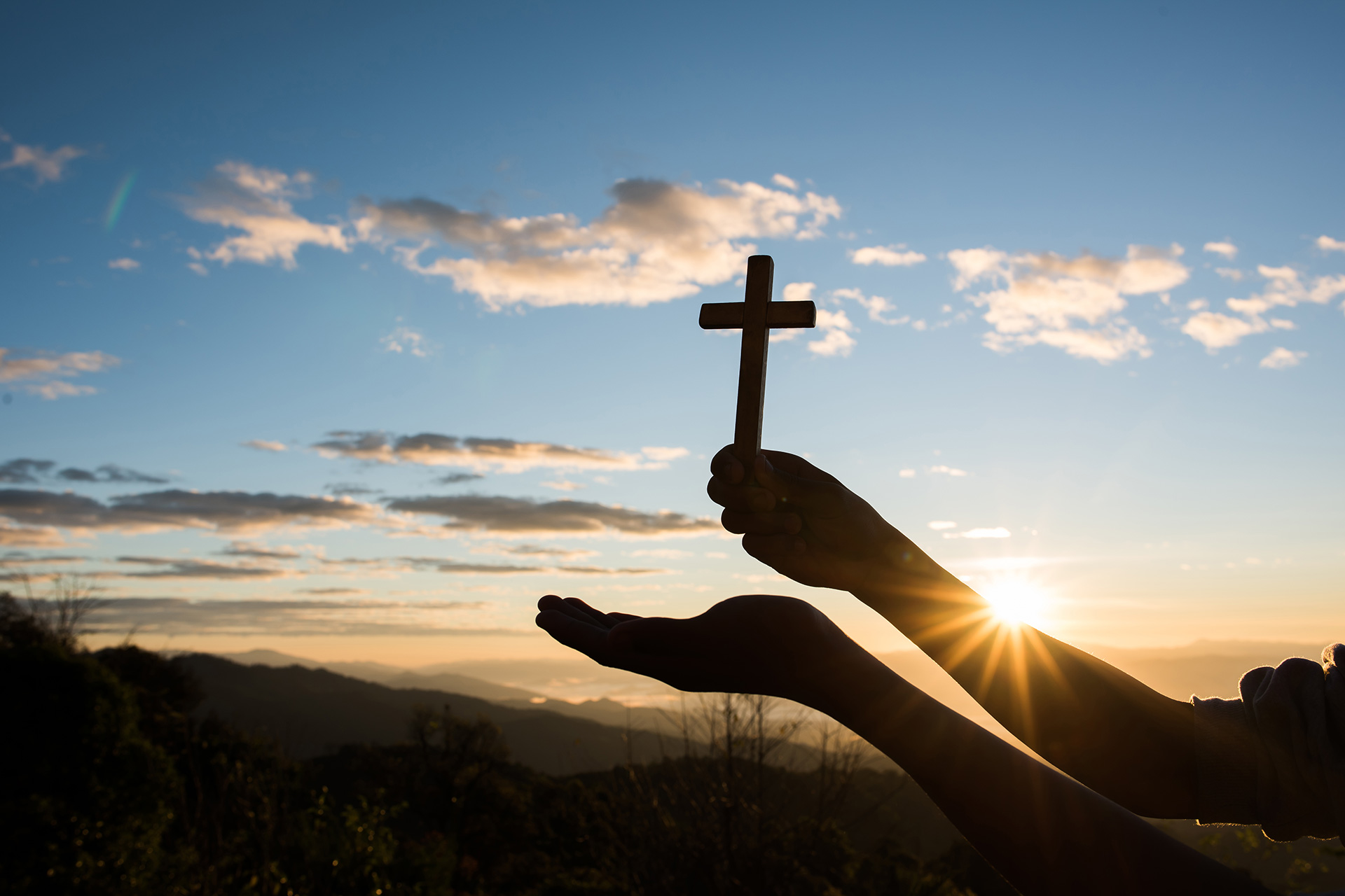 Mano levantando una cruz de madera al amanecer, símbolo de fe cristiana y espiritualidad, con un paisaje montañoso de fondo iluminado por el sol naciente.